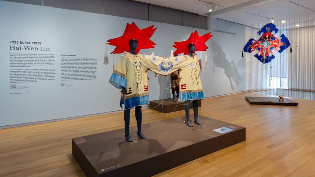 Una vista de una galería de museo que presenta la obra del ganador del Premio Burke 2025, Hai-Wen Lin. En el centro, sobre una plataforma baja, un maniquí viste una prenda artística de color crema con motivos azules, con grandes formas rojas abstractas adheridas a la parte superior que recuerdan a cometas. A la derecha, otra pieza cuelga del techo, y en la pared izquierda hay un panel explicativo que detalla el premio y la declaración del artista.