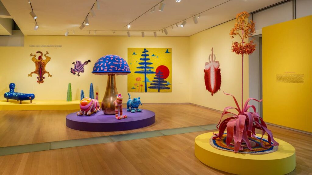 Una galería colorida y caprichosa que exhibe obras de los Haas Brothers. La habitación tiene paredes amarillas brillantes. En el centro, sobre una plataforma morada, hay esculturas coloridas y texturizadas que incluyen una seta gigante y figuras zoomorfas. En primer plano a la derecha, sobre una plataforma amarilla, se encuentra una escultura alta y compleja que recuerda a una planta suculenta con colores rosa y naranja. En las paredes cuelgan tapices con motivos de árboles y figuras animales estilizadas.
