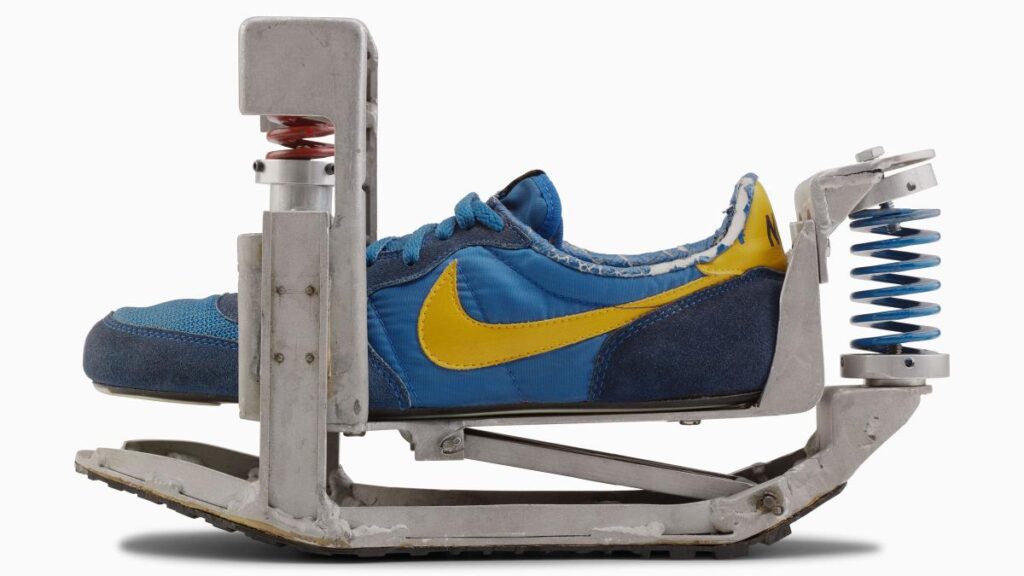 Una fotografía de estudio con fondo blanco que muestra un zapato deportivo Nike de color azul y amarillo insertado en un dispositivo mecánico experimental o de prueba. El dispositivo es una estructura de metal gris industrial que envuelve la suela y el talón del zapato, utilizando resortes helicoidales (uno sobre la puntera y otro detrás del talón) conectados a una placa base, lo que sugiere un aparato diseñado para probar la compresión, el rebote o la durabilidad de la zapatilla bajo esfuerzo mecánico.