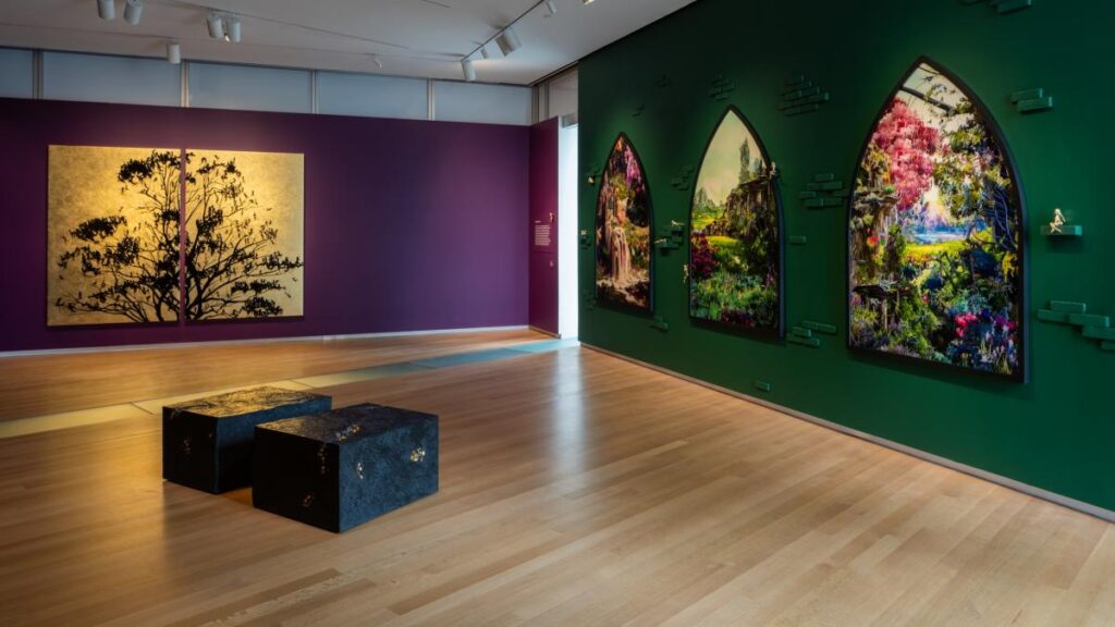 Interior de una galería de arte con paredes divididas en morado y verde oscuro. En la pared morada a la izquierda, hay dos paneles de arte dorado con siluetas de árboles negros. En la pared verde a la derecha, tres marcos con forma de arco gótico exhiben imágenes vibrantes de paisajes naturales fantásticos. En primer plano, sobre el suelo de madera, hay dos cajas negras rectangulares con detalles dorados.